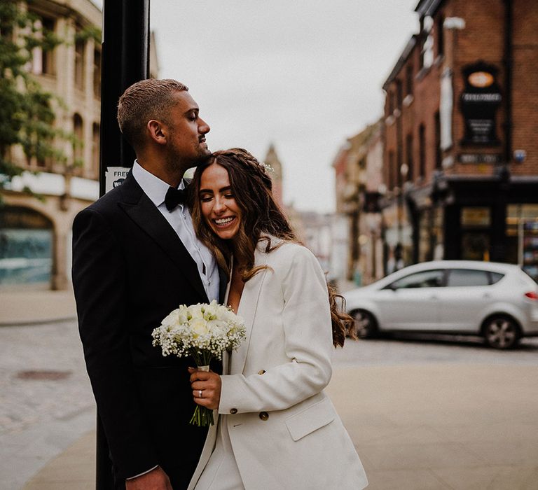 Bride in a white blazer holding a white rose and gypsophila bouquet snuggling into her groom in a black tuxedo 