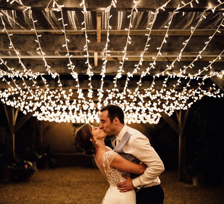 Bride in white open back Rime Arodaky wedding dress with lace train kisses groom in blue waistcoat and black trousers as they stand under a canopy of fairy lights at evening wedding reception in Dorset