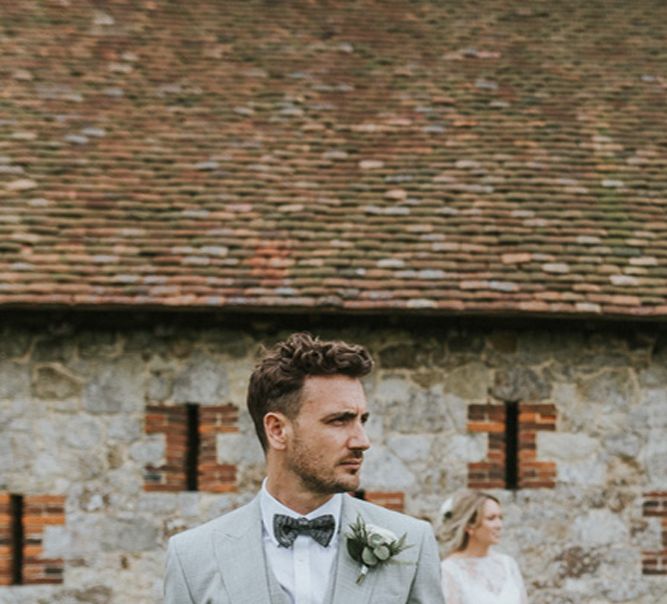 Portrait of the groom in a pale grey wedding suit and matching waistcoat with bow tie with his bride in the background