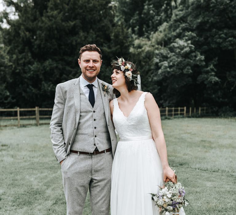 Groom in a pale grey three-piece wool wedding suit with black tie embracing his bride in a whimsical dress and flower crown 