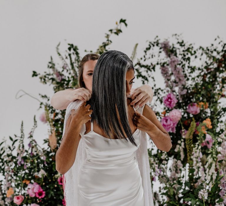 Bride in satin Halfpenny London wedding dress stands whilst wedding guest helps her put on Halfpenny London bridal cape in front of multicoloured floral installation before wedding at Loft Studios London