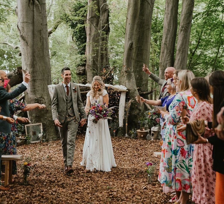 Bride in lace cap sleeved wedding dress holding mixed bridal bouquet holds hands with room in grey three piece herringbone suit walking back down the aisle during woodland wedding ceremony as guests throw confetti at late summer wedding in Norfolk