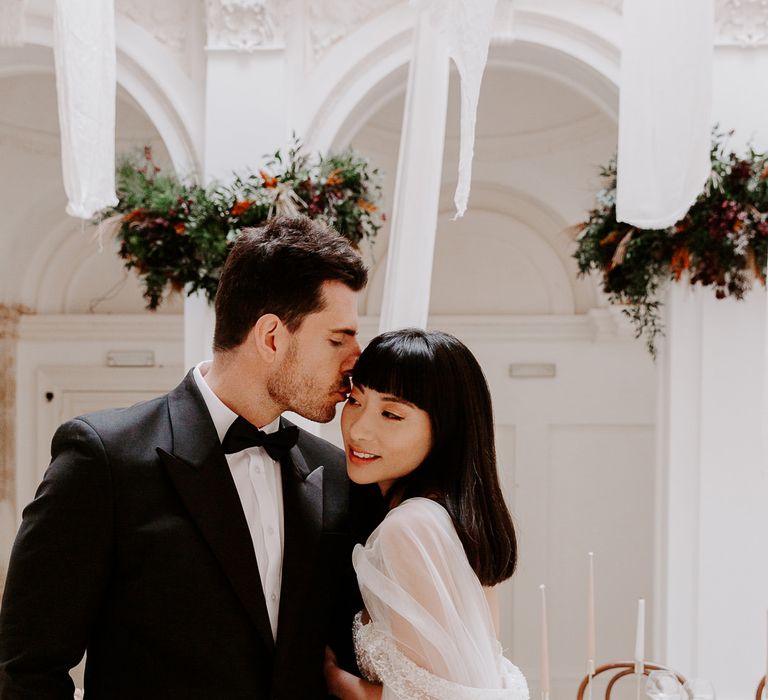 Groom in a tuxedo kissing his brides head with a blunt fringe, shoulder length hair and natural makeup