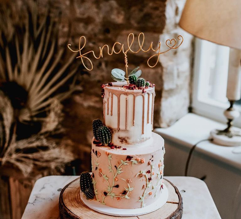 two tier drip wedding cake with flower and cactus decor and a 'finally' wire wedding cake topper 