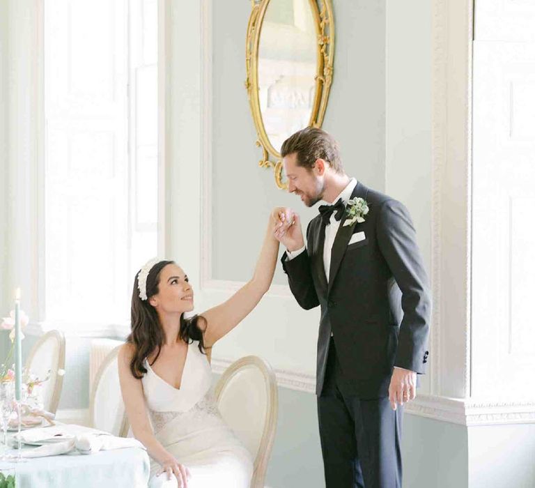 Groom in a tuxedo kissing his brides hand as she sits at the wedding reception table decorated with pastel decor and flowers 