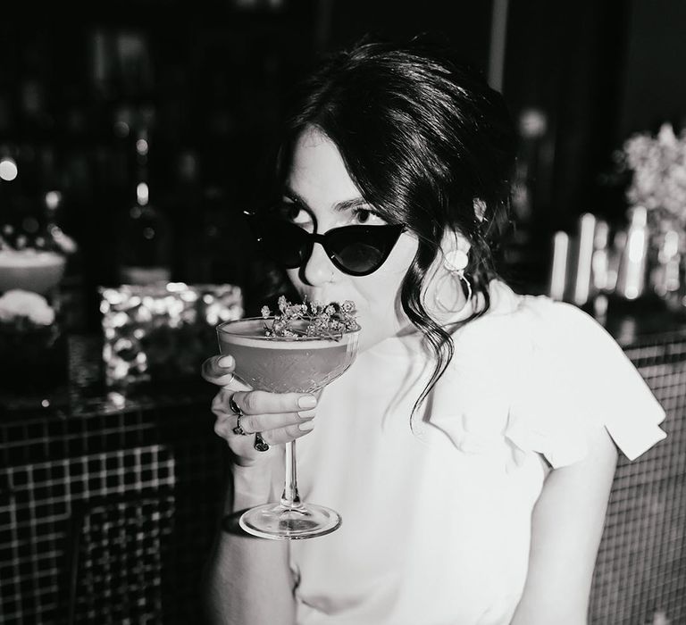 black and white picture of a stylish bride in a wedding dress with no sleeves wearing sunglasses and sipping cocktails on the bar 