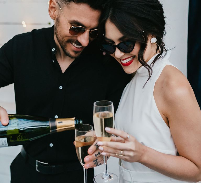 Groom in a black short sleeve shirt pouring champagne for his bride in a halterneck wedding dress with pinned up hair, sunglasses and red lipstick 