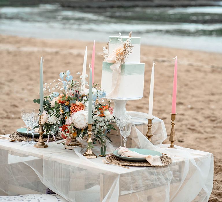 Wooden palette tablescape and cushions on the beach with pastel coloured candles and square buttercream wedding cake 