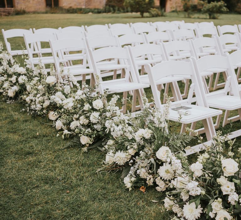 White wooden chairs with green and white floral lined aisle for outdoor wedding ceremony in Buckinghamshire