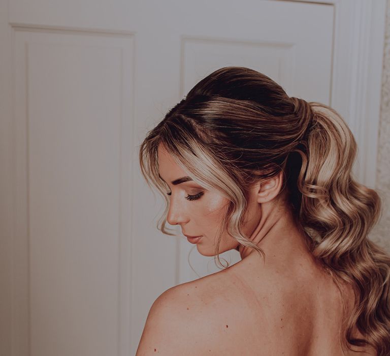 Bride looks down her shoulder as she wears her hair in a ponytail in waves