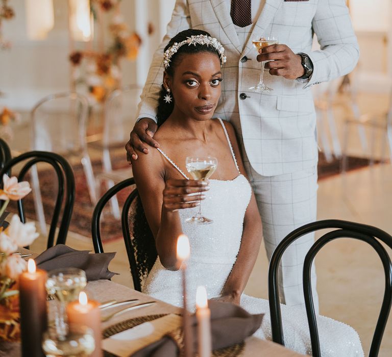 Black bride in a white sparkly wedding dress and Black groom in a beige check suit with brown tie holding champagne glasses in the air at their Kirtlington Park wedding 