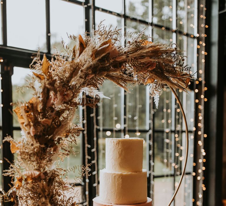 Three tier wedding cake with white and pink icing on large boho hoop cake stand with dried floral decor for rustic barn wedding