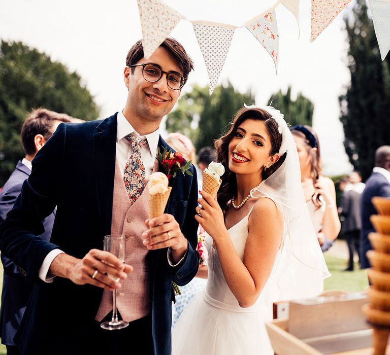 Bride in feathered headband and pearl necklace stand with groom in dark suit, baby pink waistcoat and floral tie both holding ice creams in waffle cones