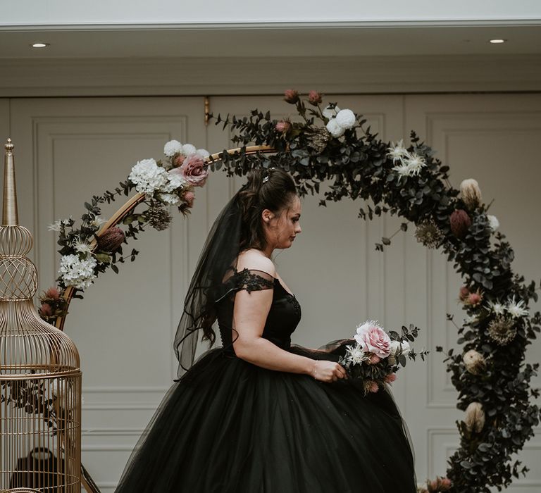 Bride wears black wedding dress whilst standing in front of floral archway complete with pastel florals 