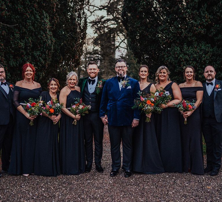 Wedding party all pose together with grooms in colourful suit jackets and bridesmaids holding colourful and bright bouquets