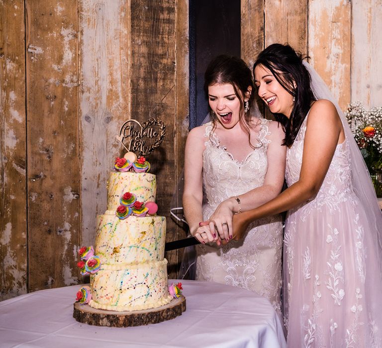 Brides cut their white frosted rainbow paint splattered wedding cake with rainbow iced roses, macarons and personalised wooden heart topper