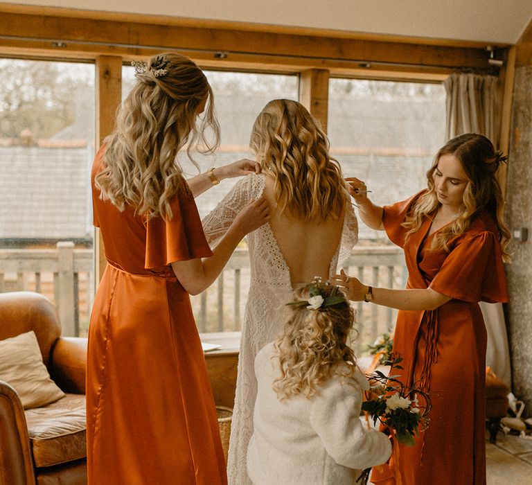 Bridesmaids in burnt orange satin wrap dresses help bride into lace gown with flower girl in white fur jacket and sparkly dress