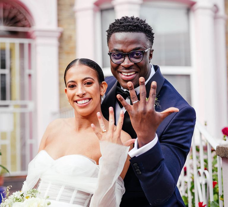 Bride and groom lift up their hands to show off their wedding bands in their outfits for industrial city wedding