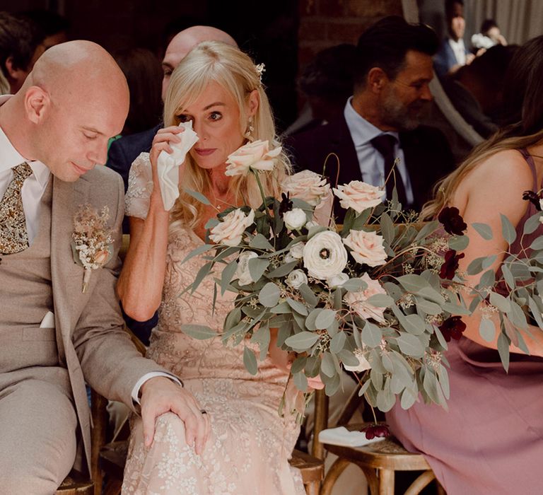 Parents wipe their eyes during emotional wedding ceremony holding pink, white and deep red wedding bouquet wearing gold and beige