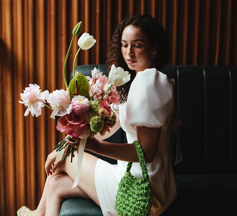 Stylish bride with naturally curly hair and freckles in a short wedding dress with green handbag and sandals holding a pink and white wedding bouquet 