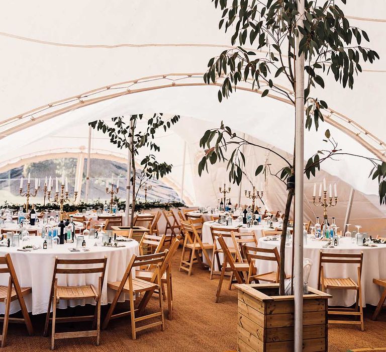 Marquee with circular tables with white wooden chairs and candelabra centrepieces with greenery 