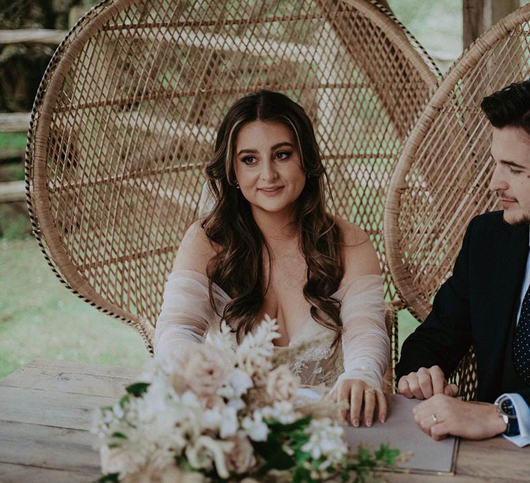 Bride and groom sit in two peacock chairs as they sign the register 