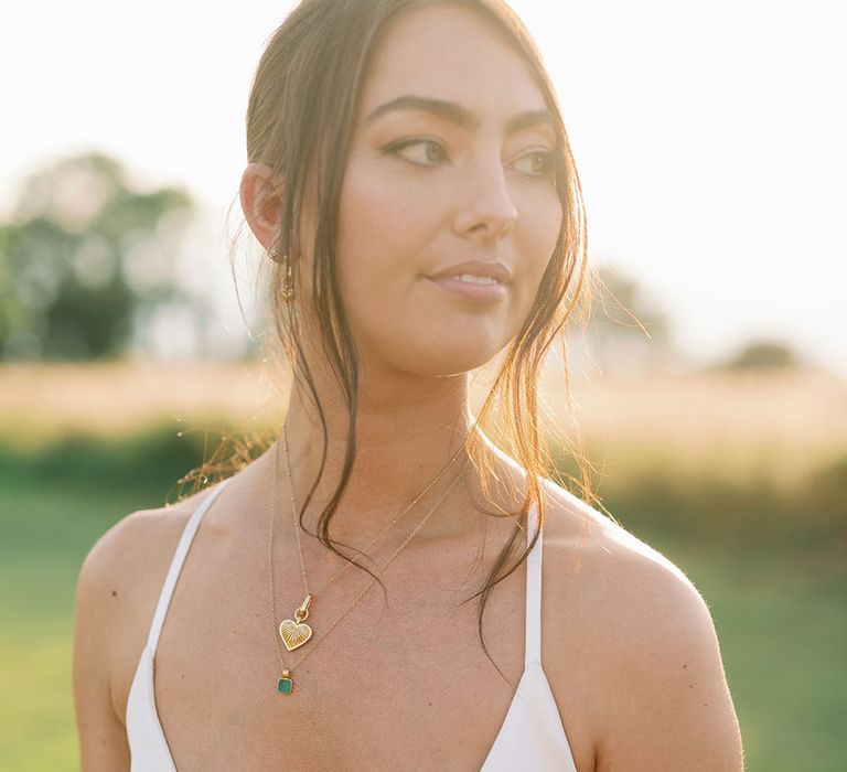 Bride wears natural makeup and low cut DIY wedding gown complete with gold necklaces and brown hair pulled into low ponytail 