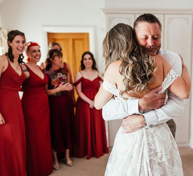 Bride in off the shoulder boho lace wedding dress hugs her father with the bridesmaids in the background in orange dresses