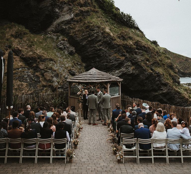 Wedding guests sit outdoors at Tunnels Beach in Devon as florals line the aisle and beach hut sits to the front