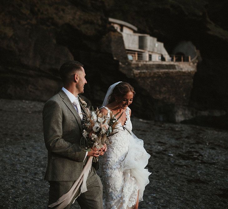 Groom holds champagne and rust coloured bridal bouquet as he walks along the beach with his bride for couples portraits 