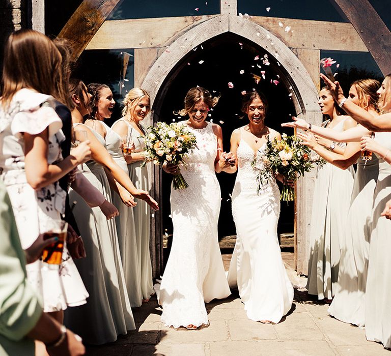 Bridesmaids in soft green dresses and other wedding guests throw confetti over the brides