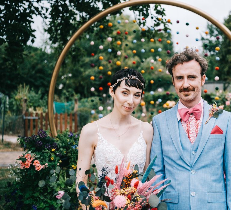 Bride holds bright dried floral bouquet and stands with her groom in dusty blue suit with pink 70s styled ruffled shirt in front of pom pom wedding decorations