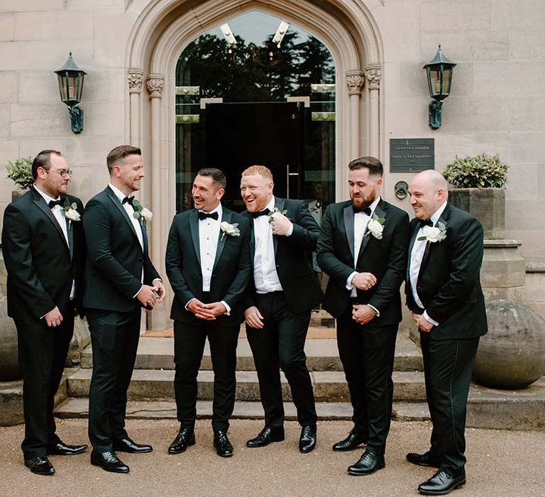 Groom stands with his groomsmen who wear black tie and white rose buttonholes 