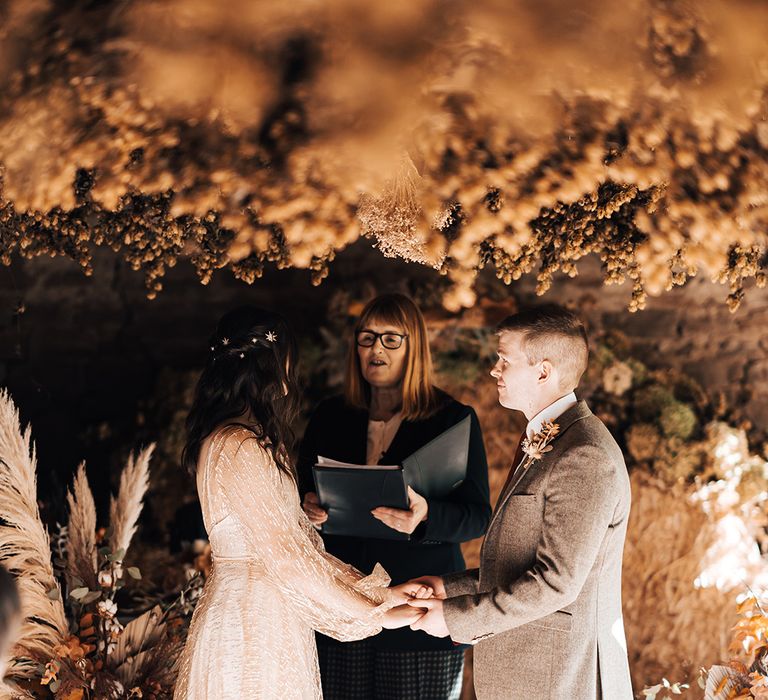 Bride in sheer gold wedding dress during wedding ceremony with her groom surrounded by dried florals at Lyde Court