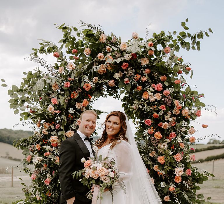 Bride in sheer full sleeved wedding dress stands with her groom in front of orange floral archway outdoors at Botley Hill Barn