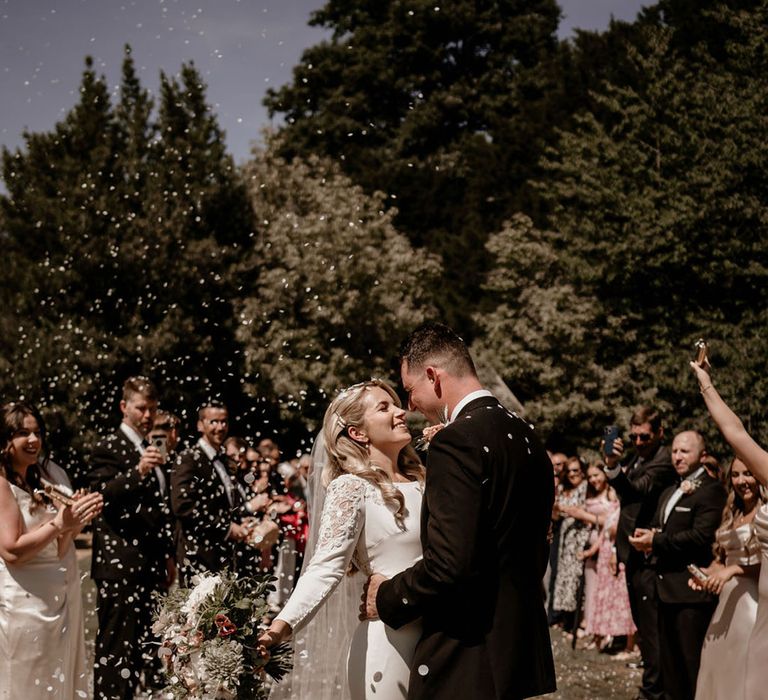 Bride in a long sleeve wedding dress with intricate detail on the sleeves standing with the groom in black tie smiling under white confetti 