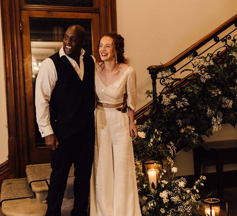 Groom in black waistcoat and suit trousers, bride in two piece bridal outfit with sparkly crop top and flowy white wide leg trousers laughing and embracing