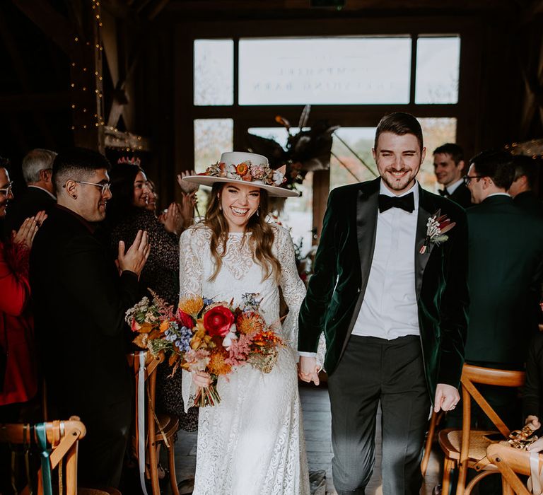 Bride wearing a white hat decorated with colourful flowers that match the bouquet walking back down the aisle with the groom in a velvet suit jacket