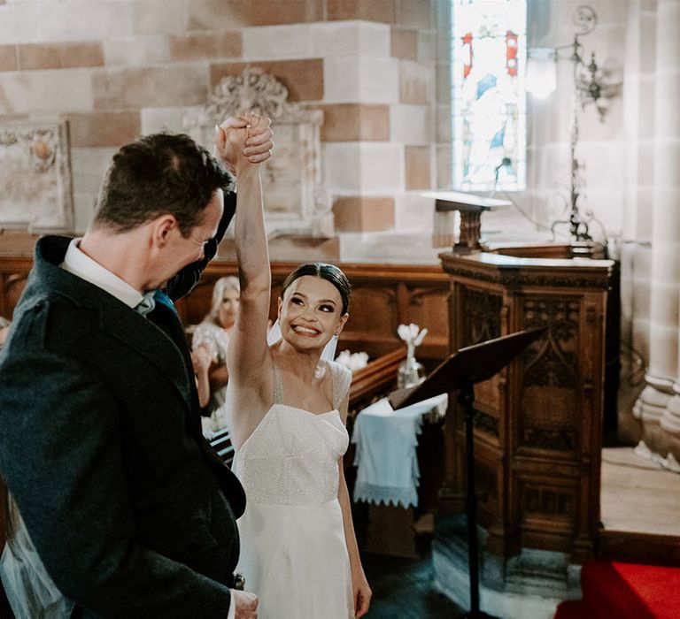 Groom lifts the bride's hand as she beams and they're announced a married couple at their church wedding 