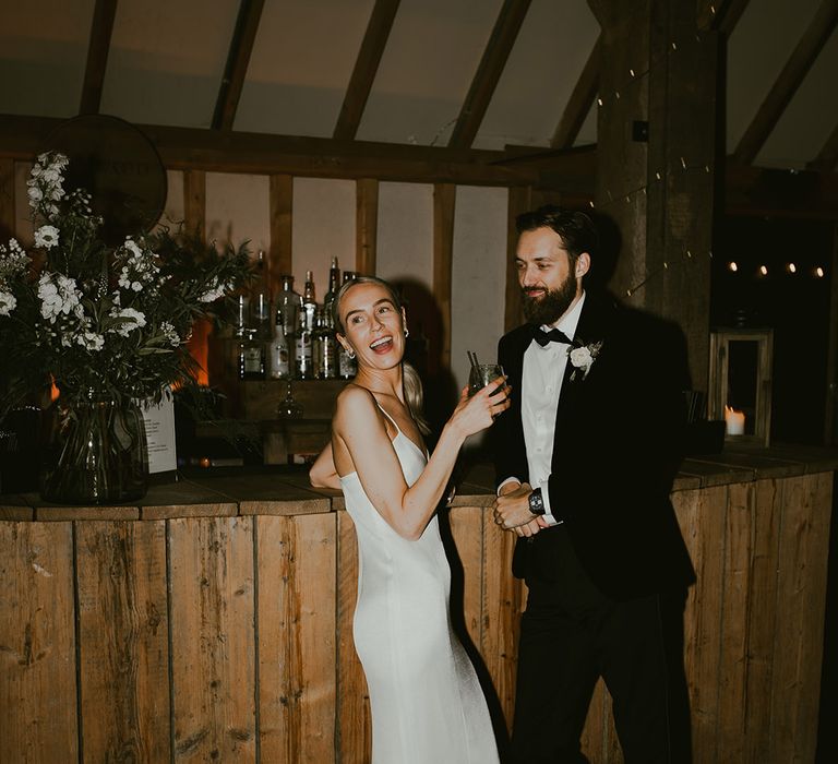 The bride enjoys cocktails from the bar with the groom at their minimal chic Southend Barns wedding 