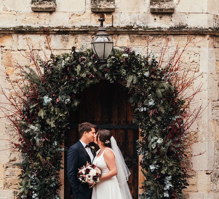 Groom in black tie kissing the bride in a white dress with a puddle train in front of the red wedding flower arch decoration 