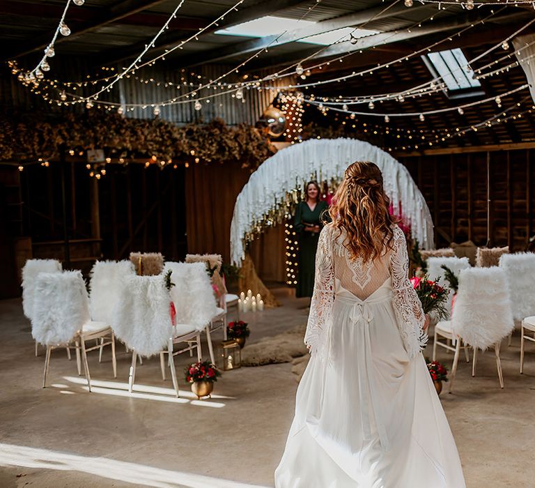 Bride in long sleeve lace wedding dress with tassels, sheer open back detail and white ribbon tie walking down the aisle with fairy lights, disco ball wedding decor, fluffy white rugs on the seats and large semi-circle arch decor with white, gold and hot pink streamers  