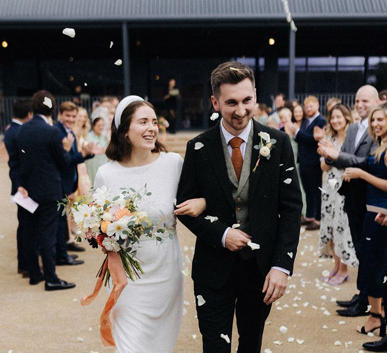 Groom in three piece suit with grey tweed waistcoat and orange tie walking with linked arms with the bride with cool headband bridal accessories as they exit to confetti 