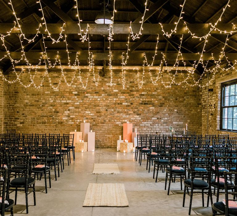 Fairy lights hang from industrial ceiling complete with pastel styled blocks and white pillar candles as black seats line the aisle 