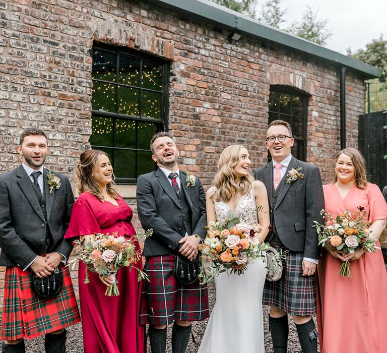 Bride & groom stand with their groomsmen in tartan kilts and brides in Rewritten Bridesmaid dresses 