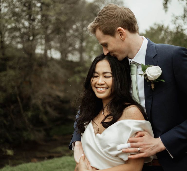 Groom in a blue suit, sage green tie and white rose buttonhole kisses the bride on the forehead wearing a Eva Lendel wedding dress 