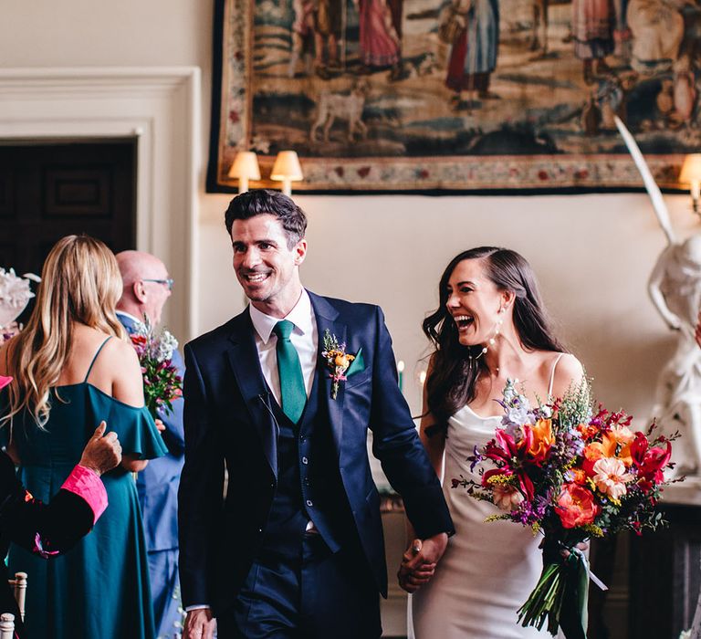 Bride with pearl earrings and satin wedding dress walks back down the aisle with the groom in a navy suit as a married couple 