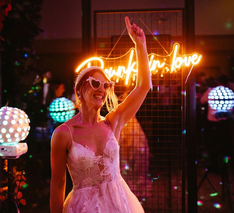 Bride wears heart shaped sunglasses and dances in front of custom neon wedding sign