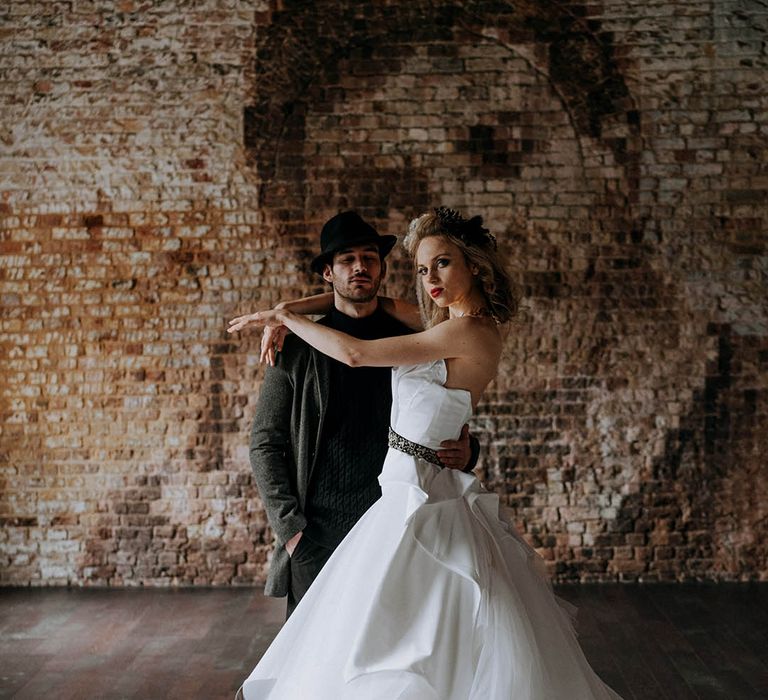 Groom in dark grey jacket, black turtle neck, black trousers and a black fedora hat embracing bride in romantic organza and tulle a-line wedding day bridal ballgown with a flowing skirt, sculpted bandeau top and patterned black belt