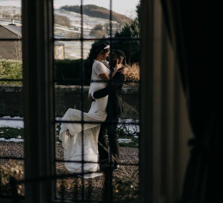 Bride in v neck short sleeve wedding dress with front slit and button detailing and groom in classic black tuxedo hugging on the grounds of Yorkshire wedding venue 